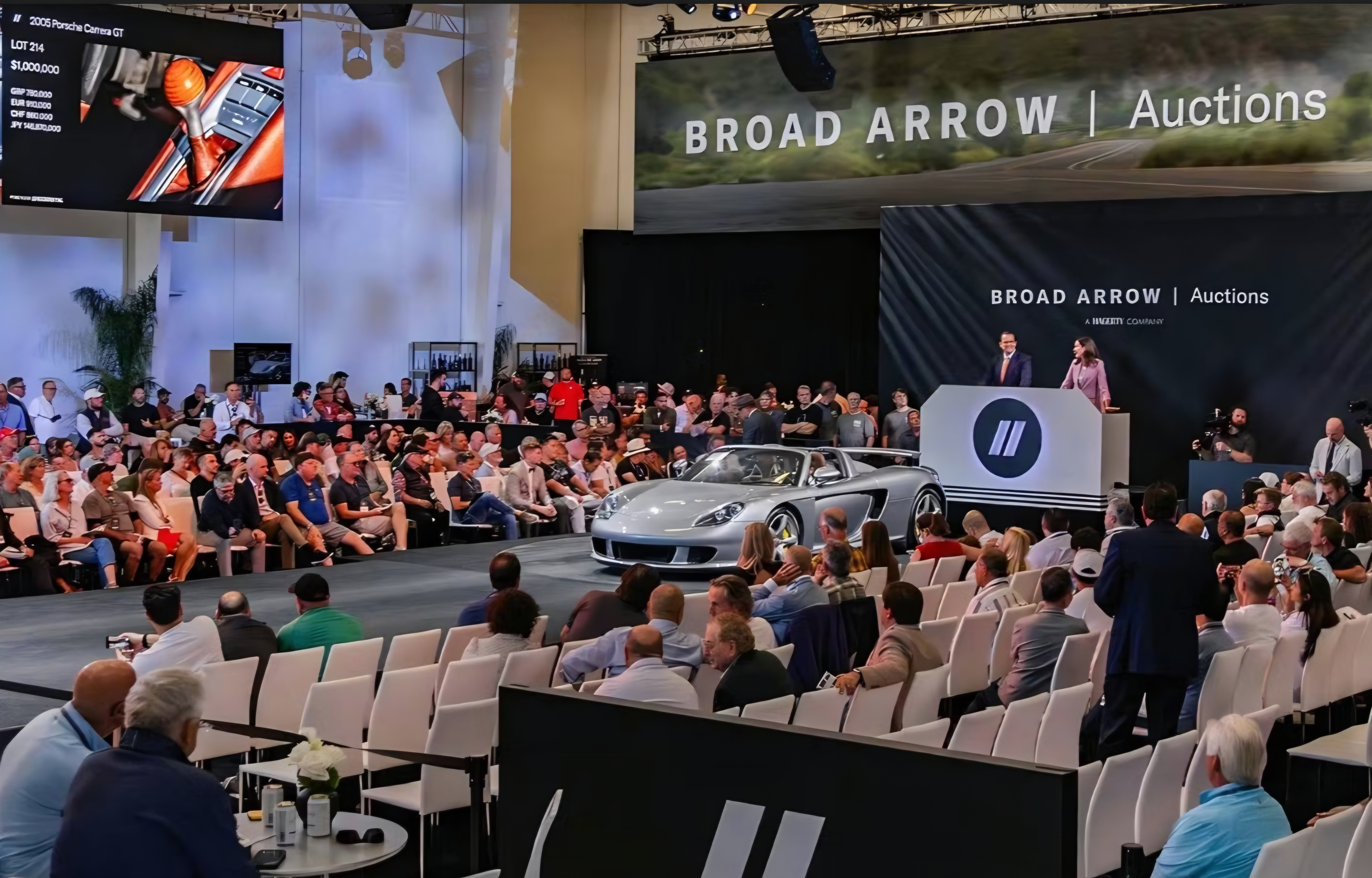 A large crowd of people gathered around a stage where a car is being auctioned off, with a prominent banner reading "BROAD...